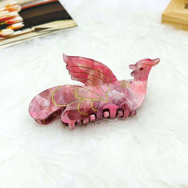 Peut inclure: Pince &agrave; cheveux rose et or en forme d'oiseau avec les ailes d&eacute;ploy&eacute;es. La pince a un motif marbr&eacute; et des accents dor&eacute;s.
