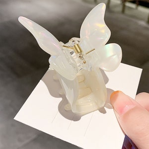 May include: A white butterfly-shaped hair clip with a gold spring mechanism. The clip is made of plastic and has a iridescent finish.
