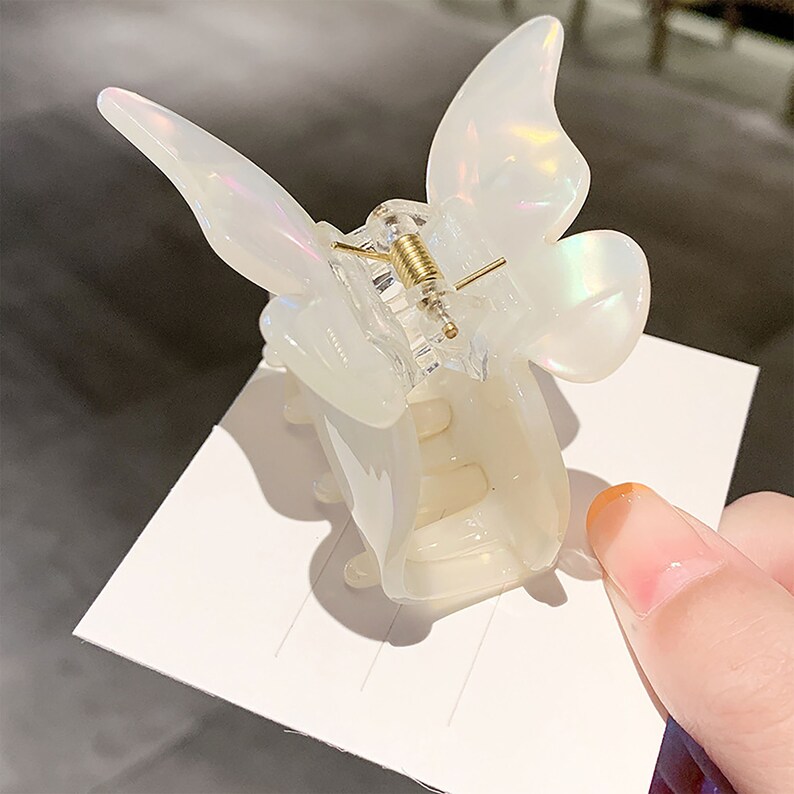 May include: A white butterfly-shaped hair clip with a gold spring mechanism. The clip is made of plastic and has a iridescent finish.