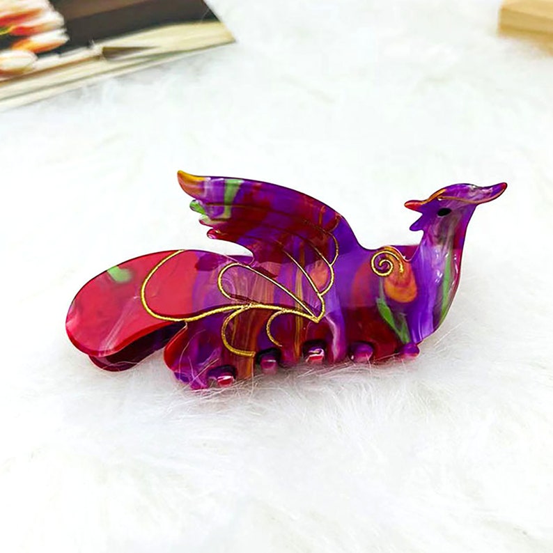 Peut inclure: Une pince &agrave; cheveux en plastique de couleur violette et rouge avec un motif de ph&eacute;nix. La pince a des accents dor&eacute;s et un motif en spirale.