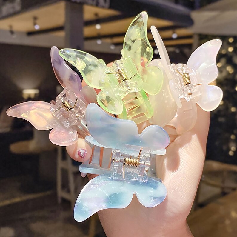 May include: Four butterfly-shaped hair clips in different pastel colors. The clips are made of clear plastic with a iridescent finish. The clips are arranged in a hand, with the largest clip in the center.