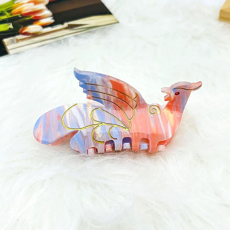 Peut inclure: Une pince &agrave; cheveux rose et bleue avec un motif dor&eacute; en forme d'oiseau aux ailes d&eacute;ploy&eacute;es. La pince a un bijou rouge sur la t&ecirc;te de l'oiseau.