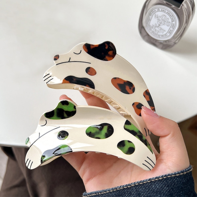 May include: Two plastic hair clips in the shape of a dog. One clip is brown and black with white spots. The other clip is white with green spots.