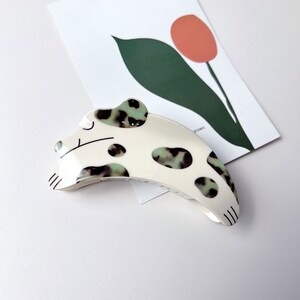 May include: White and green spotted dog-shaped hair clip. The clip is curved and has a black outline.