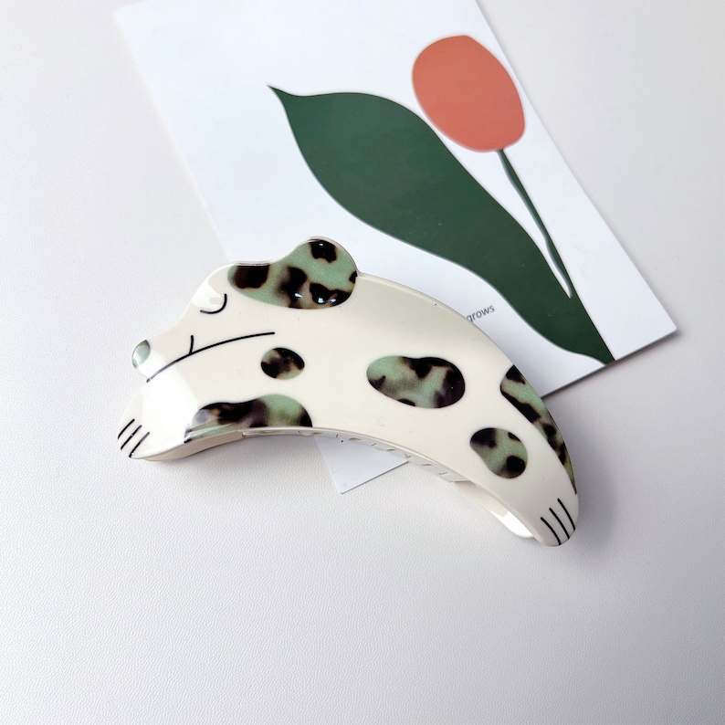 May include: White and green spotted dog-shaped hair clip. The clip is curved and has a black outline.