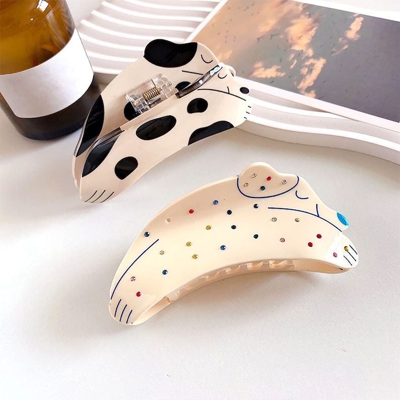 May include: Two hair clips in the shape of a dog. One clip is white with black spots and has colorful rhinestones. The other clip is black and white with a black and white spotted pattern.