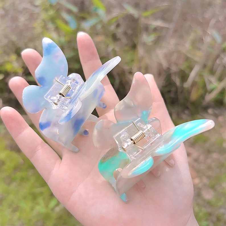 May include: Two butterfly-shaped hair clips in a translucent plastic with a marbled pattern. One clip is blue and white, the other is green and white.