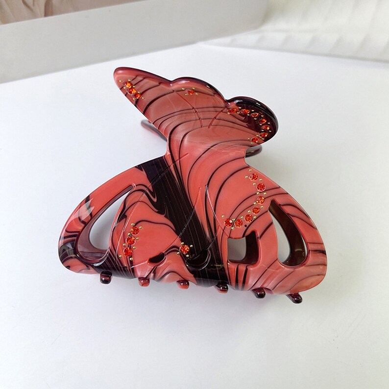 Puede incluir: Una pinza para el pelo grande de color rojo y negro con forma de mariposa con pedrer&iacute;a. La pinza est&aacute; hecha de pl&aacute;stico y tiene un patr&oacute;n de espiral.
