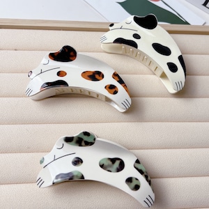 May include: Three white plastic hair clips shaped like sleeping dogs. Each clip has a different pattern of brown, black, and green spots.