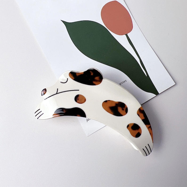 May include: White and brown spotted plastic hair clip shaped like a dog with a closed eye. The clip is curved and has a brown and white spotted pattern.