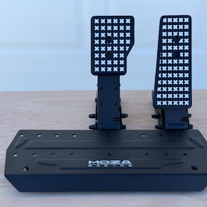 May include: Black racing pedal set with the brand name "MOZA RACING" visible. The pedals have a textured surface with a grid pattern. The set is designed for sim racing and is made of black metal.