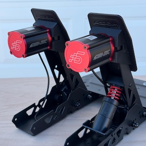 May include: A pair of black racing simulator pedals with red accents. Each pedal features a black cylinder with a red end cap and the text "SIMMAGIC". The pedals are mounted on a black base with a red spring.