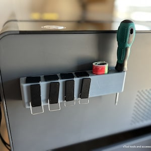 May include: A gray tool organizer with a screwdriver, measuring tape, and several black clips attached to a gray machine. The screwdriver has a green handle. The measuring tape is red and white.