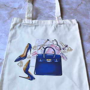 May include: A white canvas tote bag with a blue handbag, two blue high heels, gold jewelry, and a pink and white notebook with a pencil on top. The design is a colorful illustration.