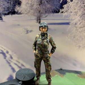 Könnte beinhalten: Eine Miniaturfigur eines Soldaten in Tarnuniform mit passender Jacke und Hose. Die Figur trägt einen dunklen Helm mit Headset und hat einen langen Zopf. Der Soldat steht mit den Händen in den Taschen auf einer grünen Oberfläche.