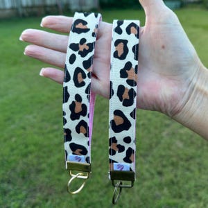 May include: Two key fobs with a cream background and a leopard print pattern in black and brown. The inside of the fob is pink. Each fob has a gold-coloured metal clasp and a key ring.