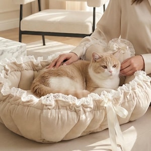Puede incluir: Una cama para gatos beige con borde con volantes y detalles de cinta. Un gato atigrado y blanco descansa dentro de la cama. La cama está sobre una otomana beige. Una mujer está sentada junto al gato, acariciándolo.
