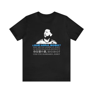 Op de afbeelding: Zwart T-shirt met een wit silhouet van een voetballer en de tekst "¿QUÉ MIRA, BOBO?" in blauw, gevolgd door vertalingen in het Engels, Portugees, Chinees en Italiaans.