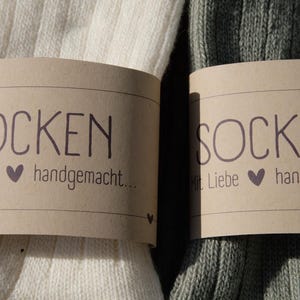 Op de afbeelding: Twee paar sokken, een wit paar en een groen paar, met bruine papieren etiketten waarop "Socken" en "Mit Liebe handgemacht..." staat.