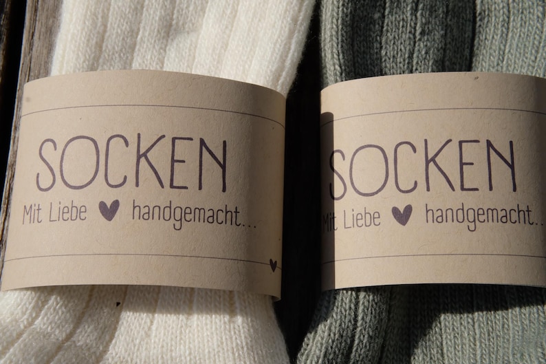 Op de afbeelding: Twee paar sokken, een wit paar en een groen paar, met bruine papieren etiketten waarop "Socken" en "Mit Liebe handgemacht..." staat.