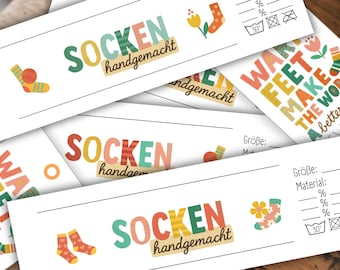 Op de afbeelding: Verschillende witte rechthoekige labels met kleurrijke tekst en sokillustraties. De labels lezen "SOCKEN handgemacht" en "WA FEET MAKE THE WORLD a better PLACE". Sommige labels bevatten wasvoorschriften.