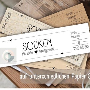 Op de afbeelding: Een wit label met zwarte tekst die "SOCKEN Mit Liebe handgemacht..." en een klein hartje-symbool zegt. Het label ligt op een stapel bruine papieren labels met zwarte tekst en symbolen. De tekst op de bruine labels omvat "Gr&ouml;&szlig;e", "Material" en percentages. De labels bevinden zich op een houten oppervlak.