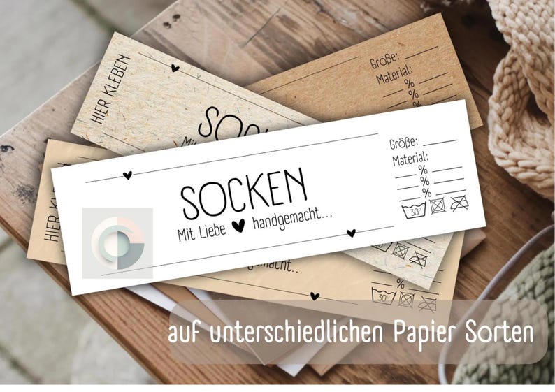 Op de afbeelding: Een wit label met zwarte tekst die "SOCKEN Mit Liebe handgemacht..." en een klein hartje-symbool zegt. Het label ligt op een stapel bruine papieren labels met zwarte tekst en symbolen. De tekst op de bruine labels omvat "Gr&ouml;&szlig;e", "Material" en percentages. De labels bevinden zich op een houten oppervlak.