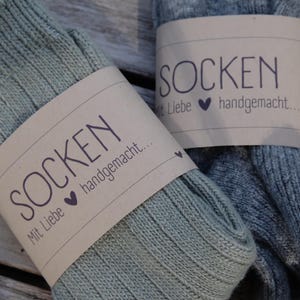 Op de afbeelding: Twee paar gebreide sokken, een groene en een grijze, met bruine papieren etiketten waarop staat "Socken mit Liebe handgemacht..." wat "Sokken met liefde handgemaakt..." betekent.