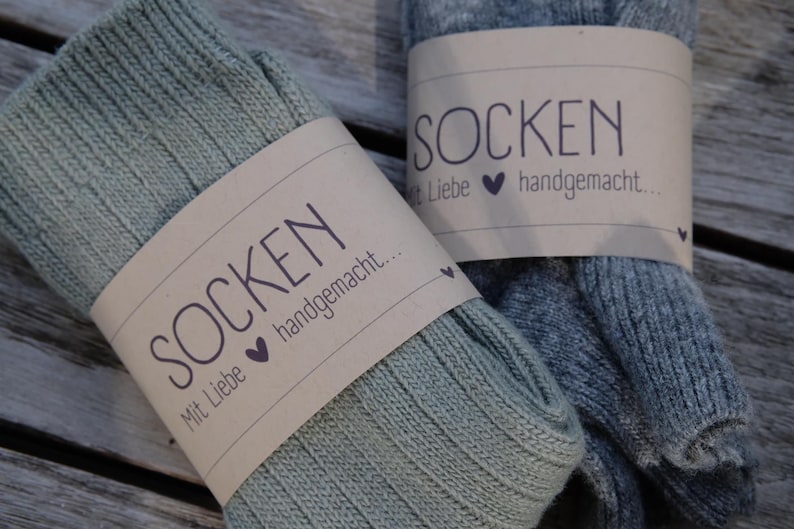 Op de afbeelding: Twee paar gebreide sokken, een groene en een grijze, met bruine papieren etiketten waarop staat "Socken mit Liebe handgemacht..." wat "Sokken met liefde handgemaakt..." betekent.