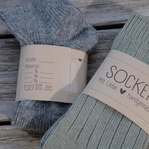 Op de afbeelding: Twee paar sokken, &eacute;&eacute;n grijs en &eacute;&eacute;n groen, met bruine papieren etiketten. Het etiket van de grijze sok heeft de tekst "Gr&ouml;&szlig;e:" en "Material:" en een wasinstructie symbool. Het etiket van de groene sok heeft de tekst "Socken handgemacht" en "Mit Liebe".