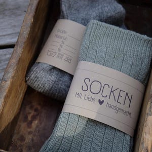 Op de afbeelding: Twee paar handgebreide sokken in een houten doos. Het ene paar is grijs en het andere is groen. De sokken zijn voorzien van een bruin etiket met de tekst "Socken Mit Liebe handgemacht."