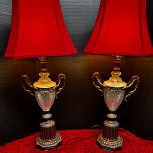 May include: Pair of vintage table lamps with red shades. The lamps feature a decorative urn-shaped base with ornate handles and a textured glass body. The bases are bronze-colored, and the shades are a vibrant red.