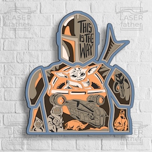 May include: A layered papercraft design of a Mandalorian character from Star Wars. The character is holding a small, green alien creature. The design includes the text "This is the way".