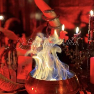 Puede incluir: Un cuenco de color cobre con llamas azules y blancas, rodeado de velas y objetos rituales. La imagen tiene un tinte rojo y el texto "Estonian Witch" es visible.