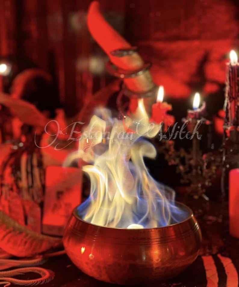 Puede incluir: Un cuenco de color cobre con llamas azules y blancas, rodeado de velas y objetos rituales. La imagen tiene un tinte rojo y el texto "Estonian Witch" es visible.