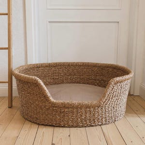 Può includere: Una cuccia rotonda per animali domestici realizzata in fibre di alghe marine intrecciate, dotata di un ingresso frontale ribassato e di un cuscino in tessuto beige chiaro che riveste la base interna, offrendo uno spazio di riposo strutturato.