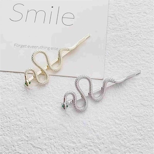 Puede incluir: Dos pinzas para el pelo en forma de serpiente, una dorada y otra plateada. Las pinzas tienen textura y forma de serpiente. El texto "Smile" y "Forget everything else" es visible en el fondo.