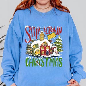 May include: A light blue crewneck sweatshirt featuring a graphic print of a snowy village scene with the red text "SMALLTOWN" and green text "CHRISTMAS". The design includes houses, a decorated pine tree, and a snowman with a candy cane.