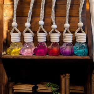May include: Eight heart-shaped glass bottles containing colourful liquids, hanging from white cords on a wooden shelf. The bottles are arranged in a row from left to right with the colours yellow, purple, pink, red, blue, green, and turquoise.
