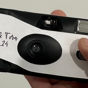 May include: A black and white disposable camera with a white label that says "Baylie & Tora 27.04.24".