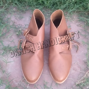 Könnte beinhalten: Ein Paar braune Lederstiefel mit Schnallen und Schnallen. Die Stiefel haben eine spitze Zehenpartie und einen niedrigen Absatz. Die Stiefel werden von Danish Handicraft hergestellt.
