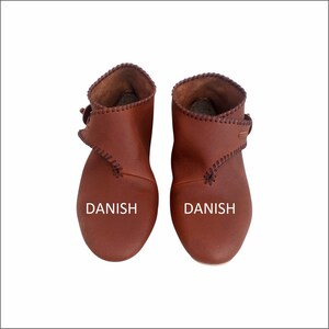 Könnte beinhalten: Ein Paar braune Lederstiefeletten mit einem Riemen und einer Schnalle. Die Stiefeletten haben eine genähte Naht am Rand und das Wort "DANISH" ist auf der Vorderseite jedes Stiefeletts aufgedruckt.