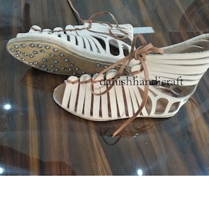 May include: White leather sandals with brown leather straps and laces. The sandals have a flat sole with metal studs.