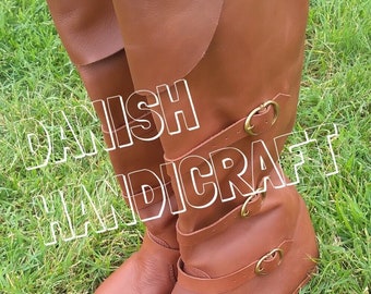 May include: A pair of brown leather boots with buckles. The boots are knee-high and have a rounded toe. The boots are on a green grassy surface.