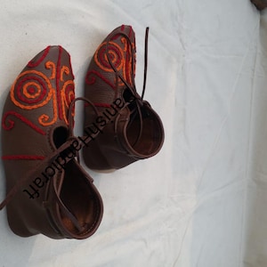 May include: A pair of brown leather shoes with orange and red embroidery. The shoes have laces and a pointed toe.
