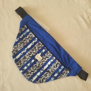 Peut inclure: Un sac banane bleu avec un tissu à motifs noirs et blancs à rayures verticales et une petite étiquette rectangulaire portant le texte NABI. Le sac possède une sangle noire réglable et une fermeture éclair sur le bord supérieur du compartiment.