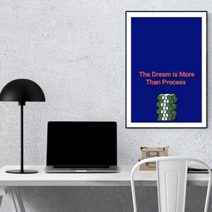 May include: A framed art print with a blue background and the text "The Dream is More Than Process" in pink. Below the text is a stack of green money. A black desk lamp, laptop, and chair are in the foreground.