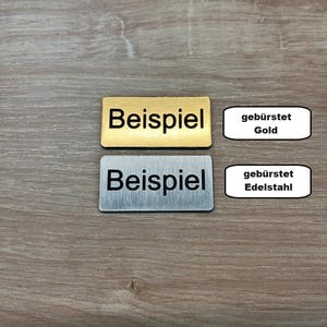 Könnte beinhalten: Zwei rechteckige Metallschilder mit dem Wort "Beispiel" in Schwarz gedruckt. Das obere Schild ist golden und das untere Schild ist silberfarben. Die Schilder befinden sich auf einer Holzoberfläche. Der Text "gebürstet Gold" ist auf einem weißen Rechteck neben dem oberen Schild gedruckt. Der Text "gebürstet Edelstahl" ist auf einem weißen Rechteck neben dem unteren Schild gedruckt.