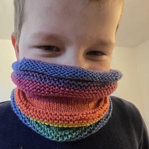 Könnte beinhalten: Ein Kind trägt einen bunten gestrickten Halsschmuck mit Streifen in Lila, Rosa, Orange, Gelb und Grün.