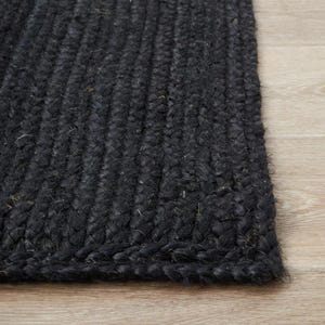 May include: A close-up of a black jute rug with a braided texture. The rug is rectangular and has a dark, charcoal color. The rug is placed on a light-colored wooden floor.
